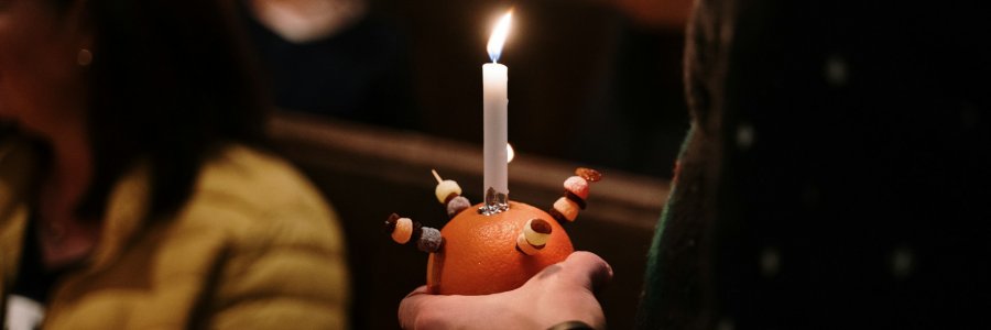 Image shows a hand holding a christingle with a lit candle.